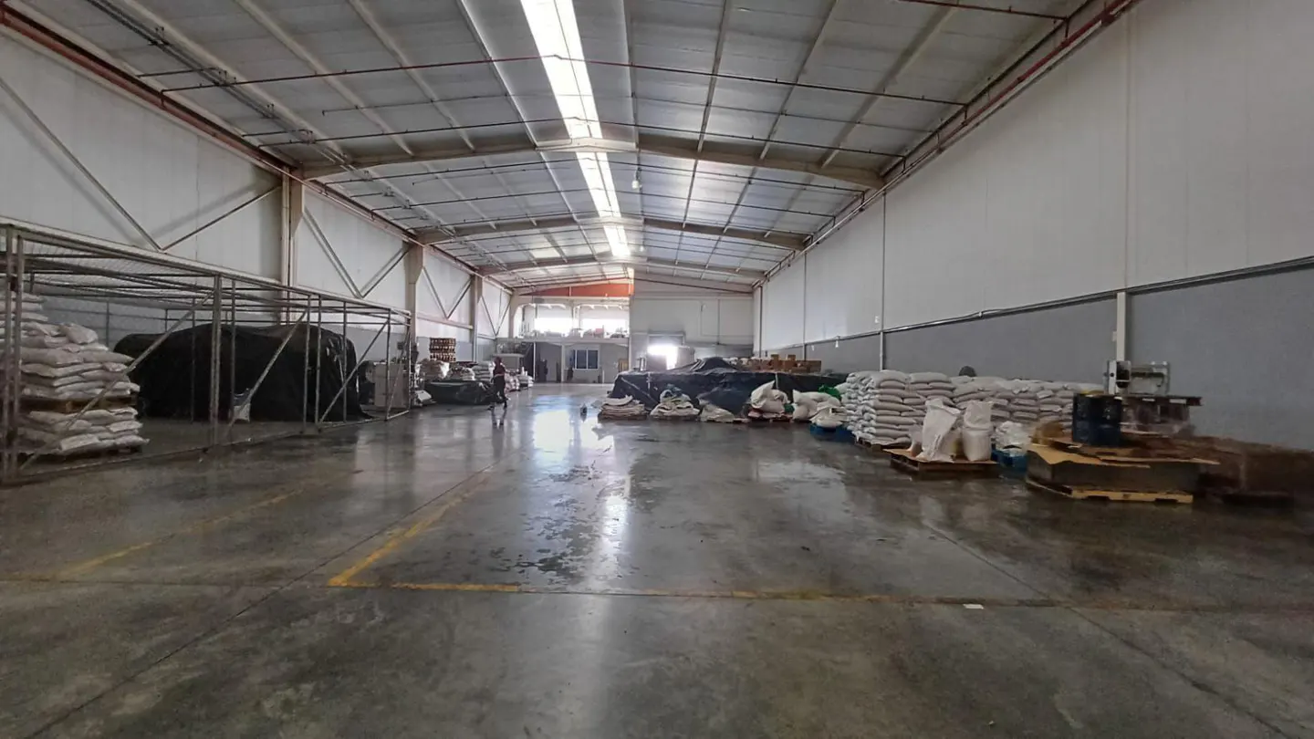 A wide shot of a large, empty warehouse with white walls and a high, silver ceiling. Bags of materials are stacked on pallets along the walls.