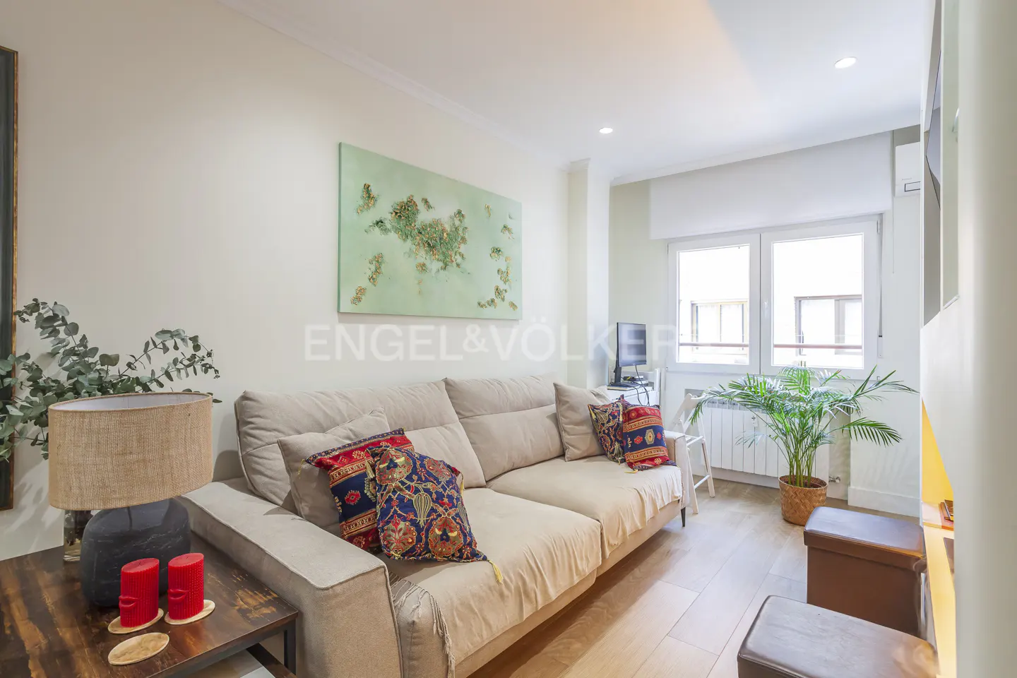 Bright living room with a beige sofa, colorful patterned pillows, abstract green wall art, and a potted plant by a window.