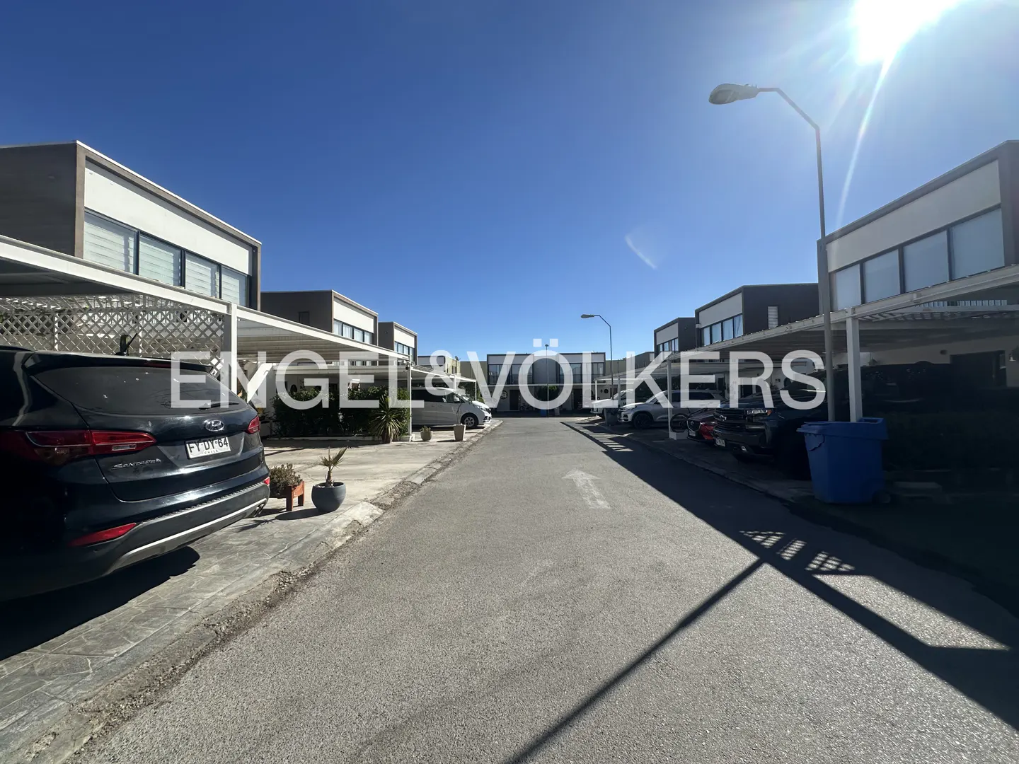 Street view of modern homes with carports under a clear blue sky. Cars are parked in front of the houses.