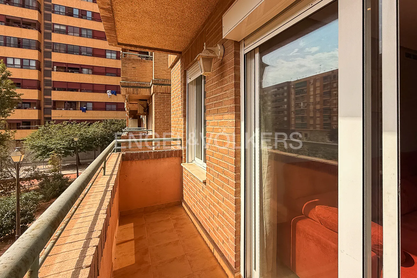 Balcony view with brick walls, metal railing, and sliding glass doors. An orange building is visible in the reflection.