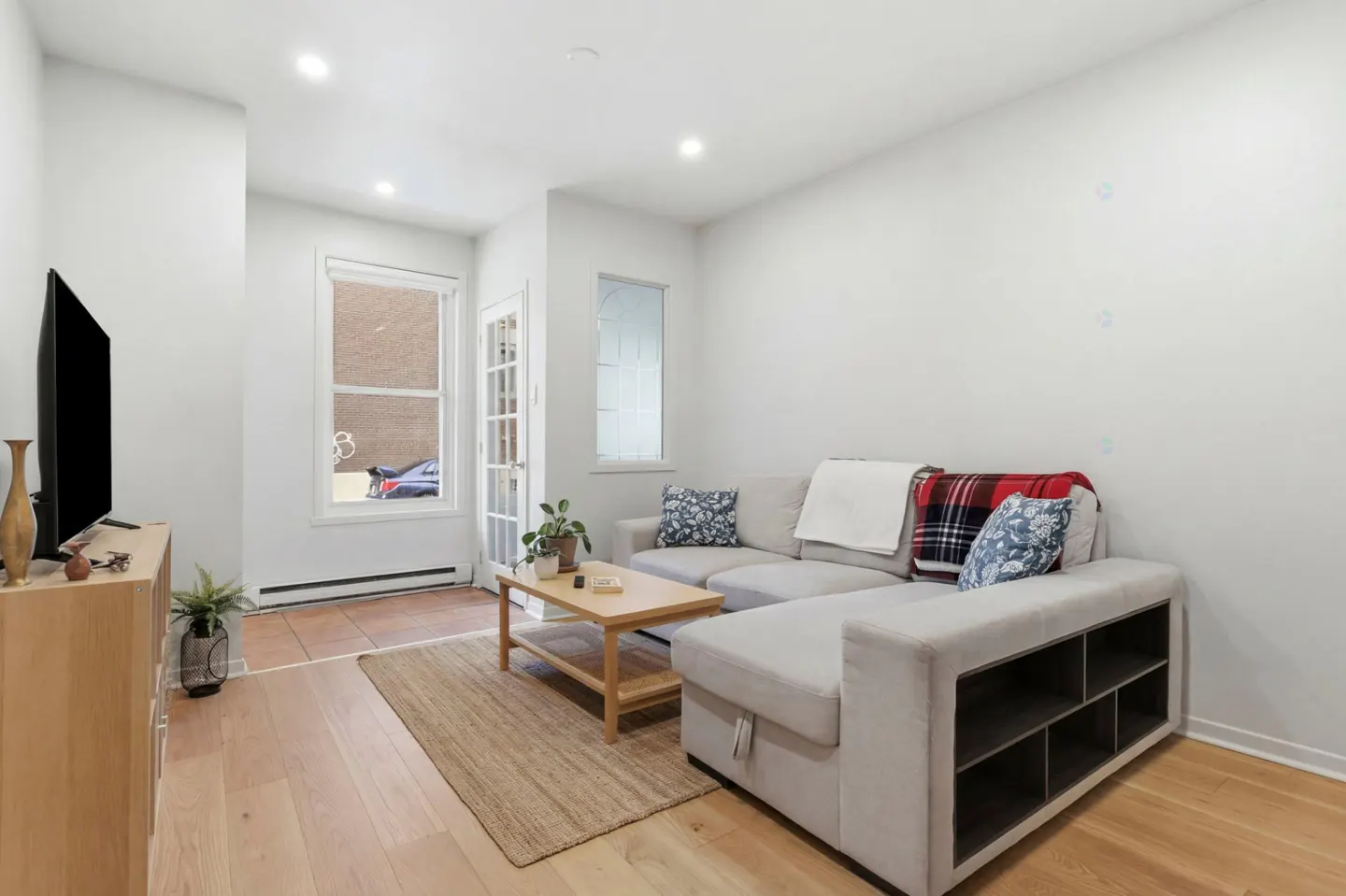 Living room with light wood floors, a gray sectional sofa with pillows, and a wooden coffee table on a jute rug. A TV sits on a wooden cabinet.