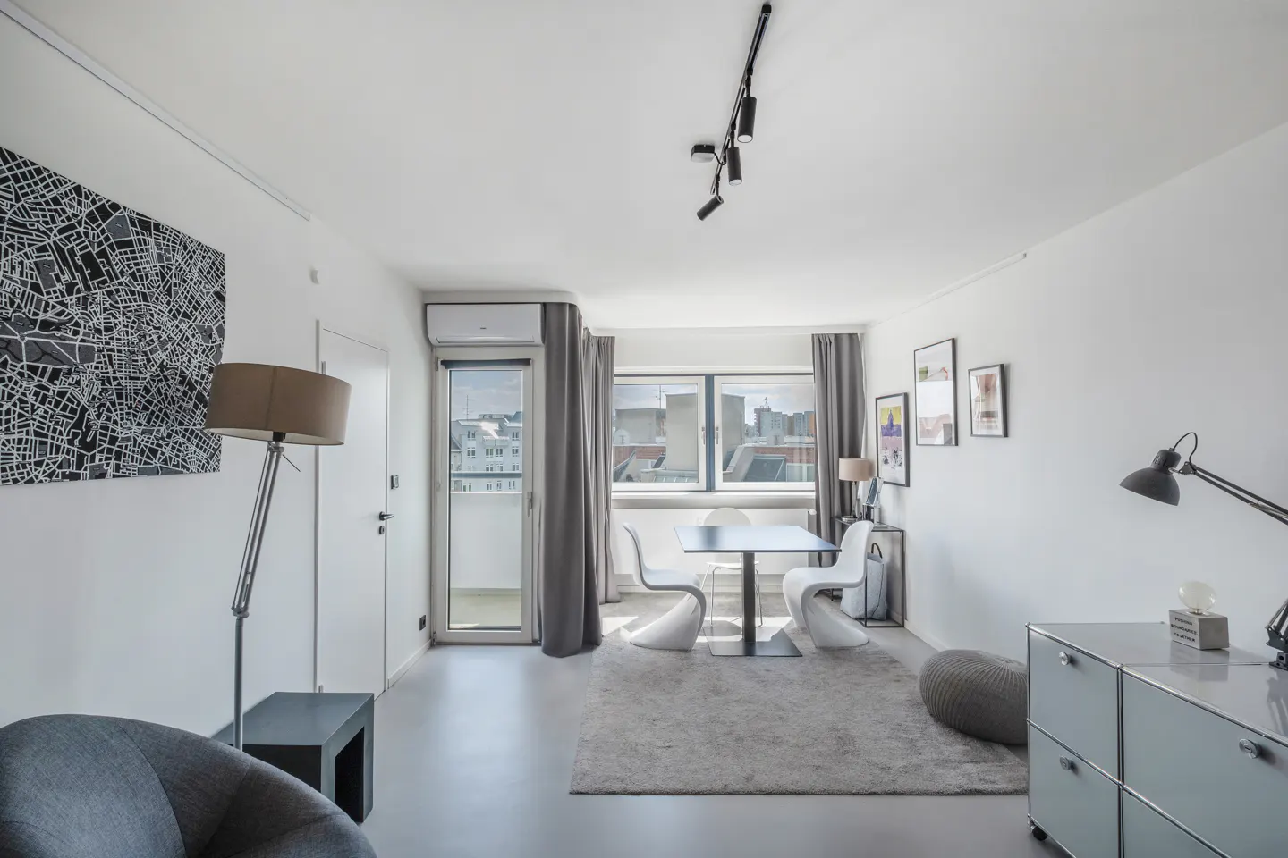 Bright, modern living room with white walls, gray rug, and city view. A black and white map art hangs on the wall. Table and chairs are near the window.