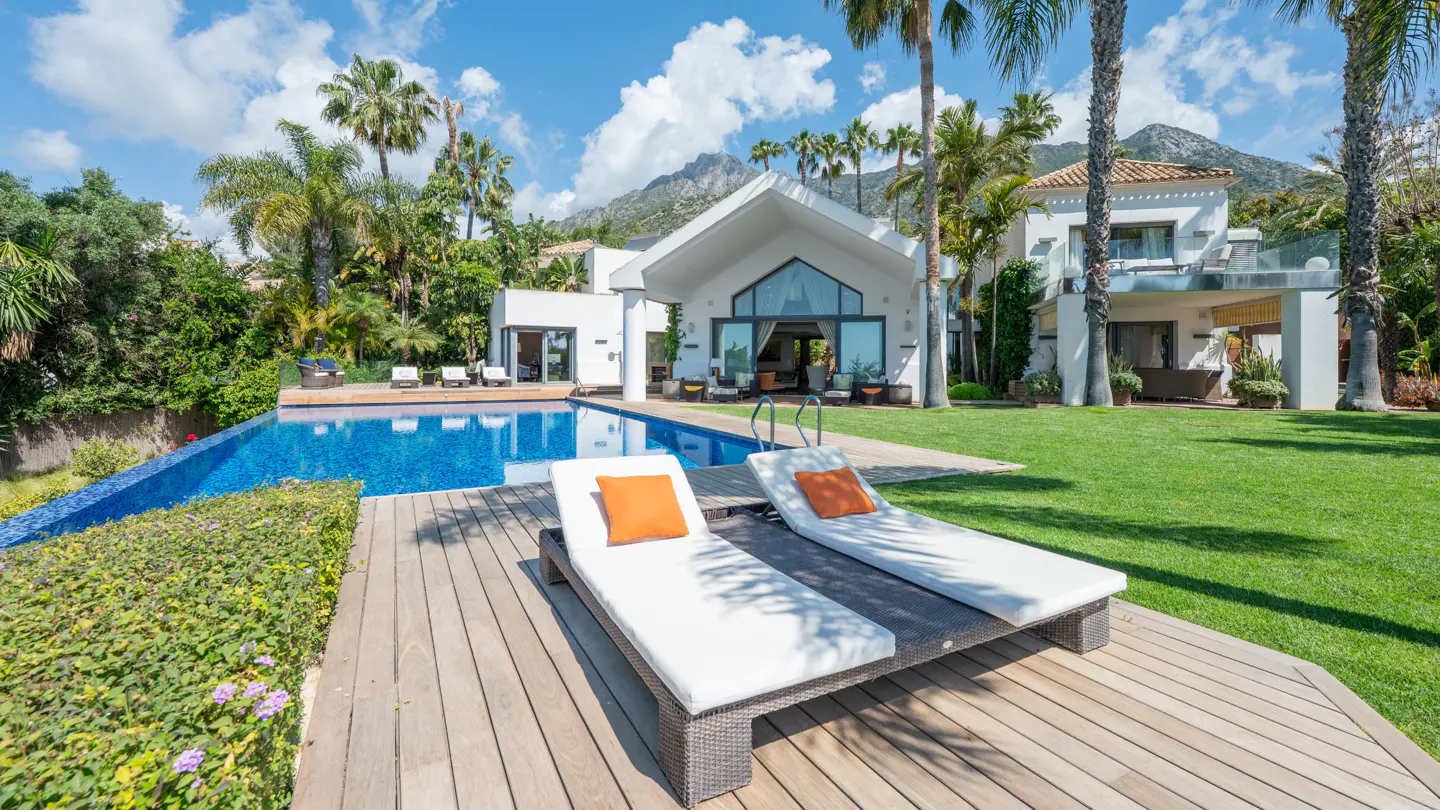 Luxury home exterior with pool, lawn, and palm trees. Two white lounge chairs with orange pillows sit on a wooden deck.