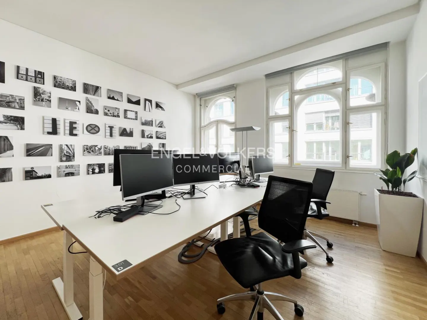 Bright office space with white walls, wood floors, and large windows. Desks with computers and black chairs are arranged in the room. Black and white photos decorate the wall.
