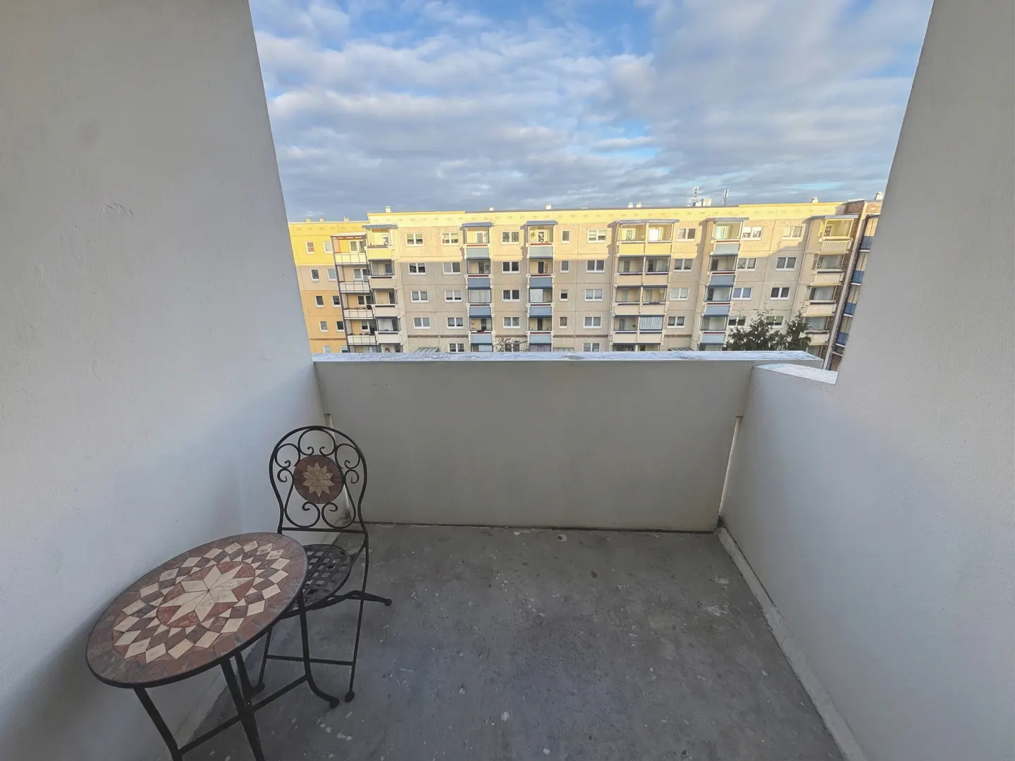 Balcony view with a round mosaic table and wrought iron chair. A large building is visible in the background under a cloudy sky.