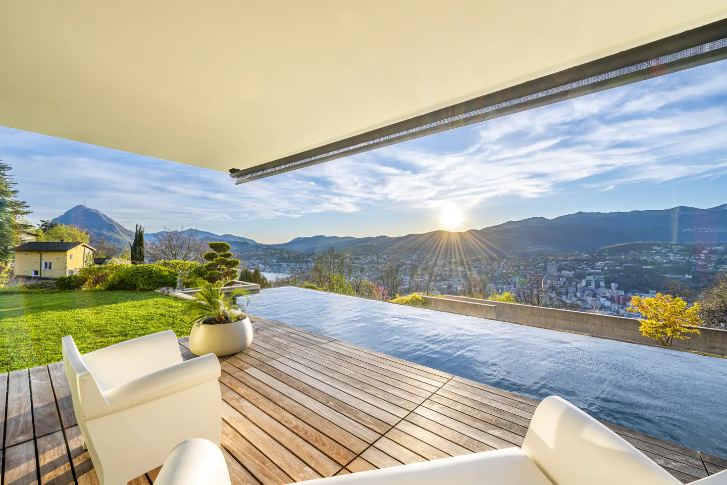 Infinity pool on a wooden deck with white chairs overlooking a mountain range at sunset.