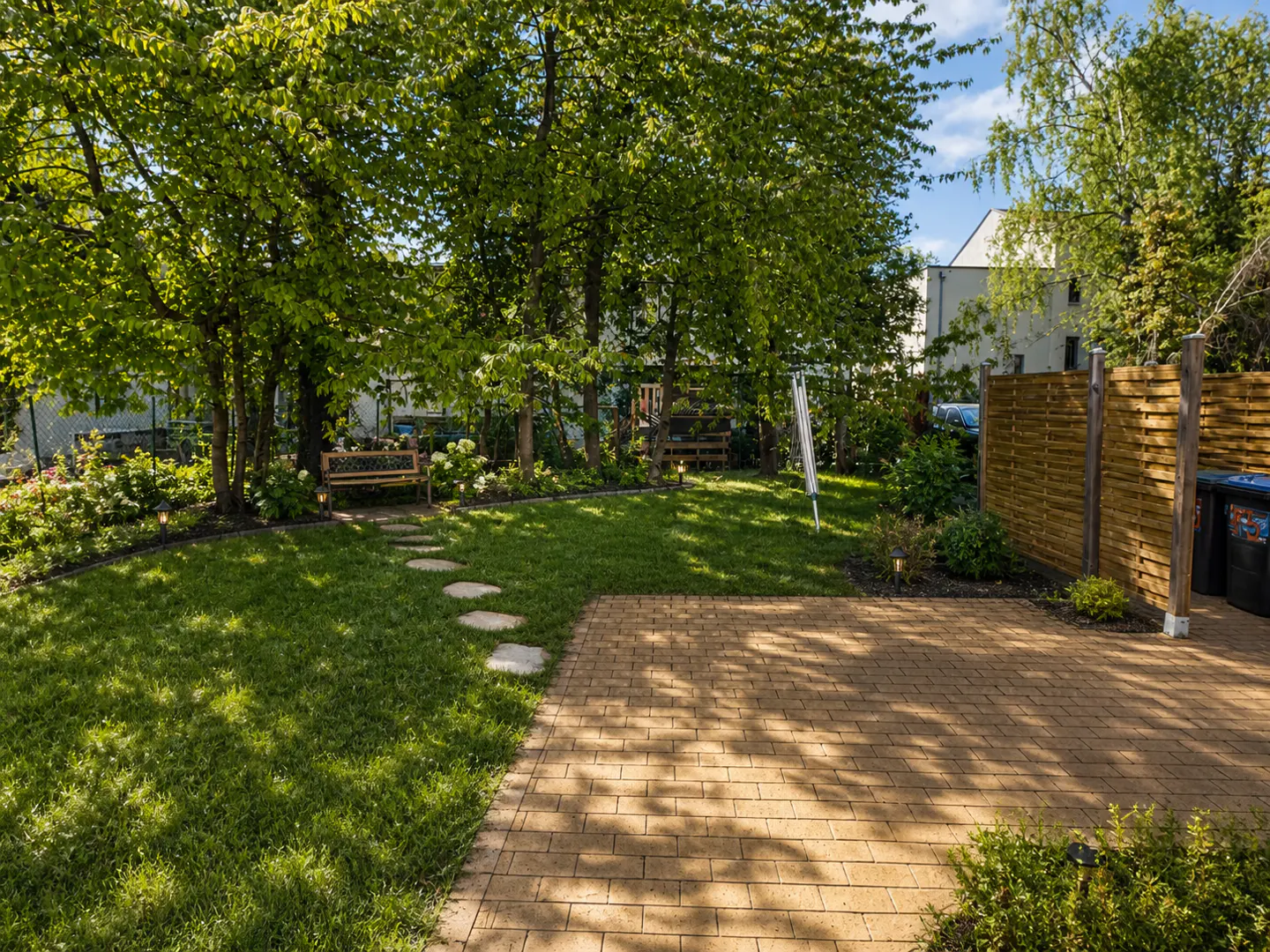 A sunny backyard with a paved patio, a stone path through a green lawn, a wooden bench, and a privacy fence.