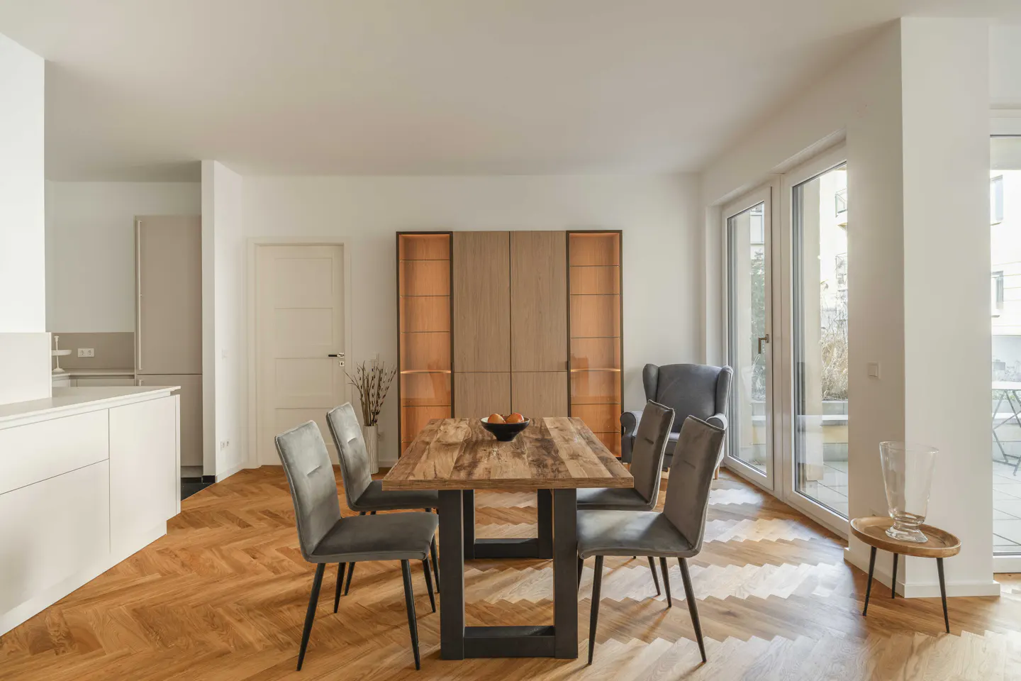 Bright dining room with wood floors, table, and gray chairs. A cabinet with glass doors and a balcony are visible.