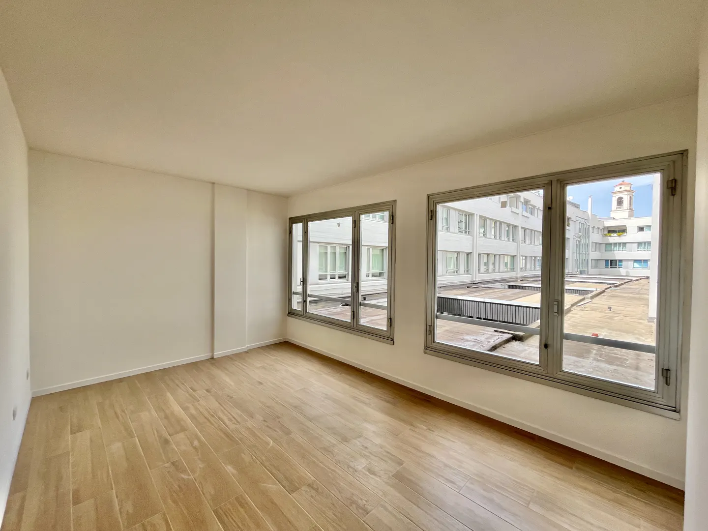 Empty room with light wood floors, white walls, and large windows. Outside, a building with a clock tower is visible.