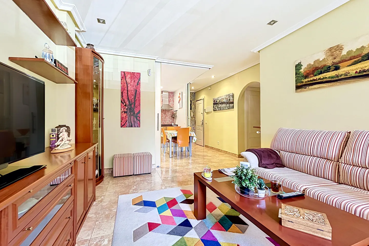 Living room with striped sofa, wood table, and colorful rug. A TV and cabinet are on the left, with a view into the dining room.