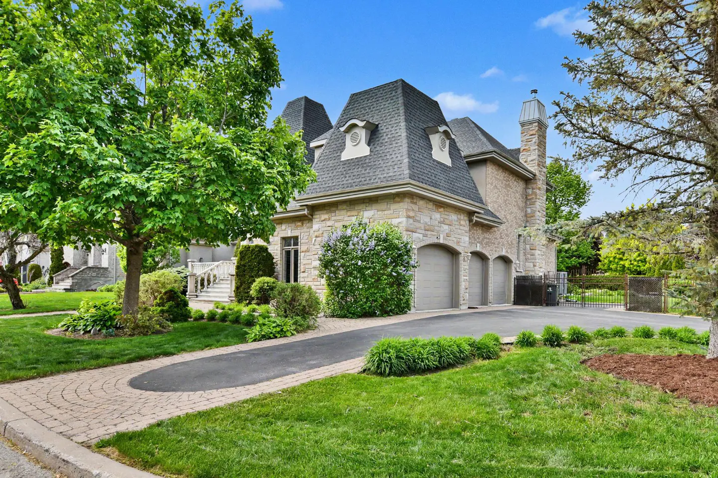 A stone house with a gray roof and three garage doors sits on a green lawn under a blue sky.