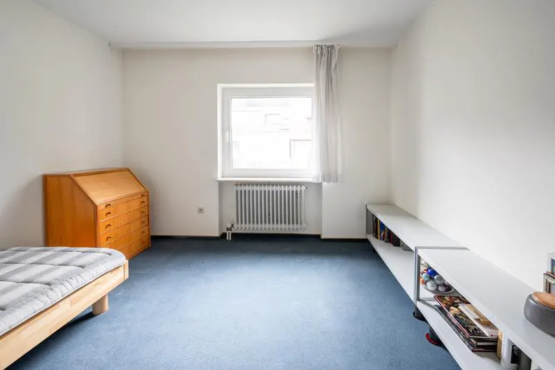 Bright room with blue carpet, white walls, and a window. A wooden desk, bed, and white shelving unit furnish the space.
