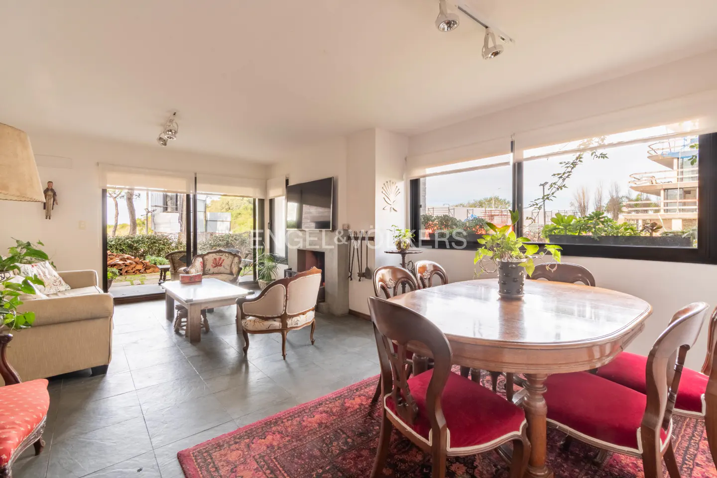 Living and dining area with a wooden table, red chairs, sofa, and large windows overlooking greenery.