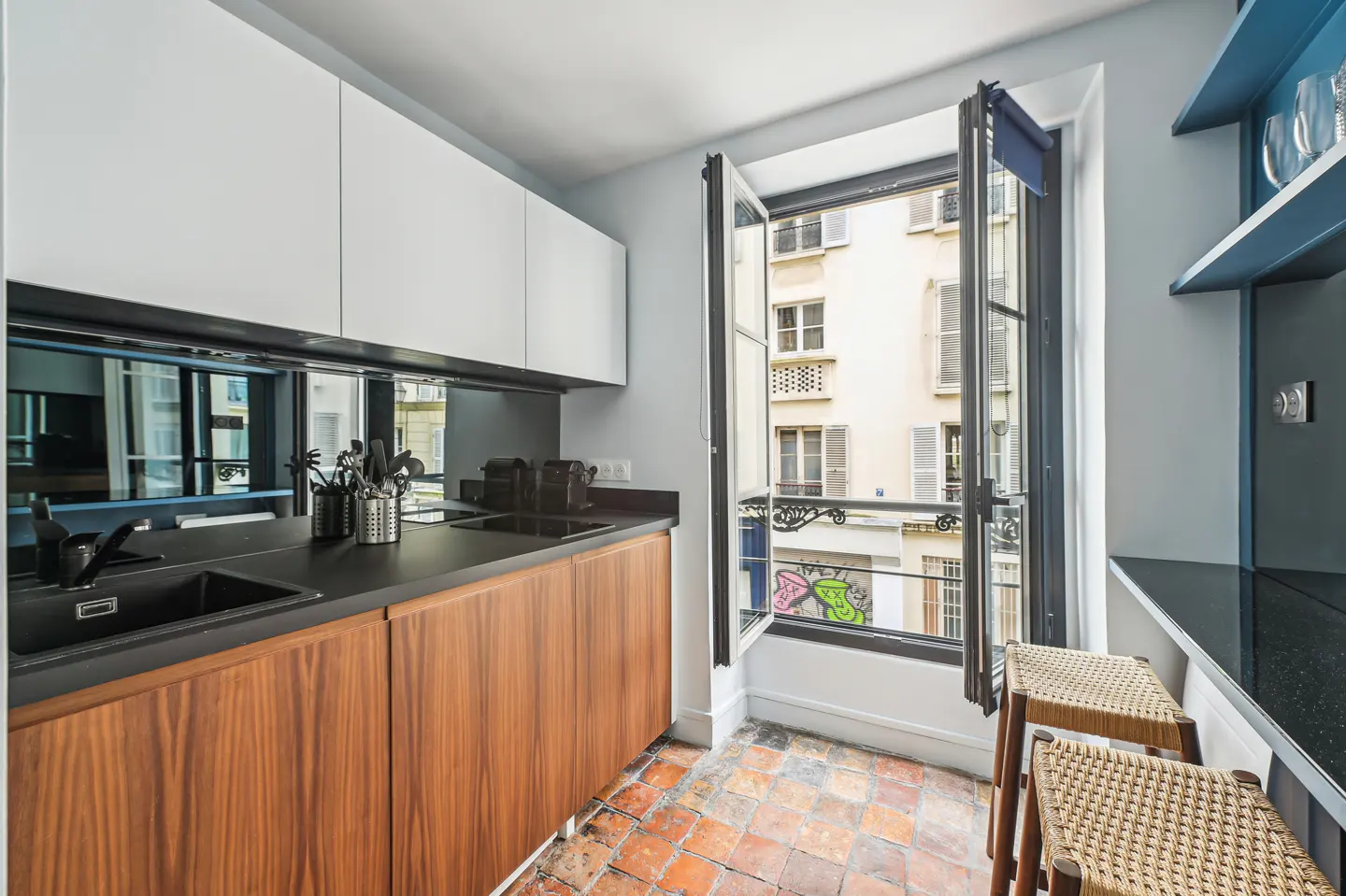 A compact kitchen with white cabinets, wood-grain lower cabinets, and a black countertop. An open window overlooks a city street. Two stools sit near a blue shelf.