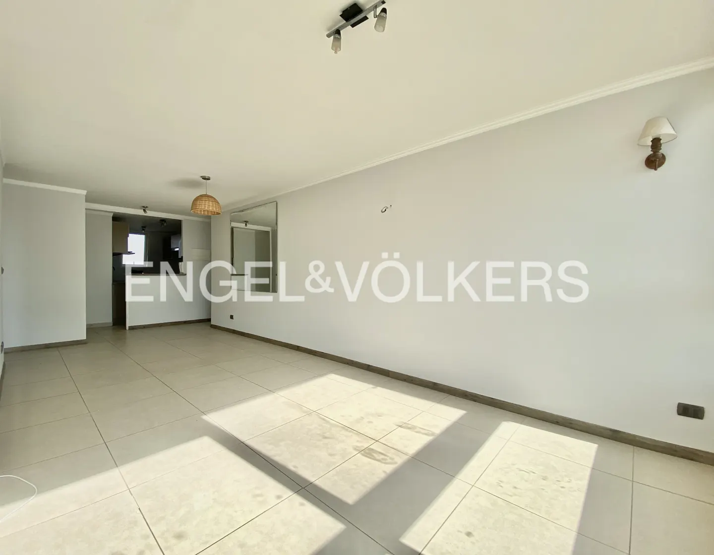 An empty living room with white walls and tile floors, featuring a kitchen area in the background.