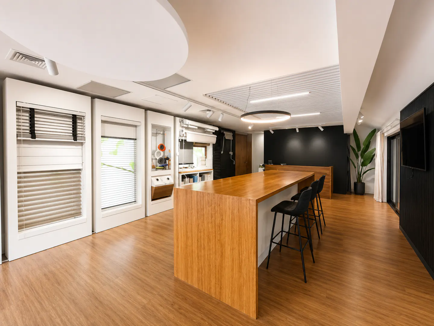 Modern showroom with wood flooring, a large wooden island with black stools, and wall displays of various window blinds.