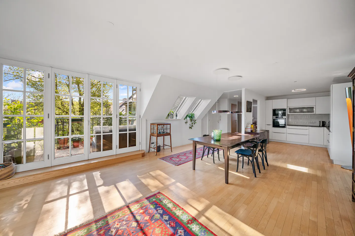 Bright, open-plan apartment with wood floors, white walls, and a modern kitchen. French doors lead to a balcony with green trees.