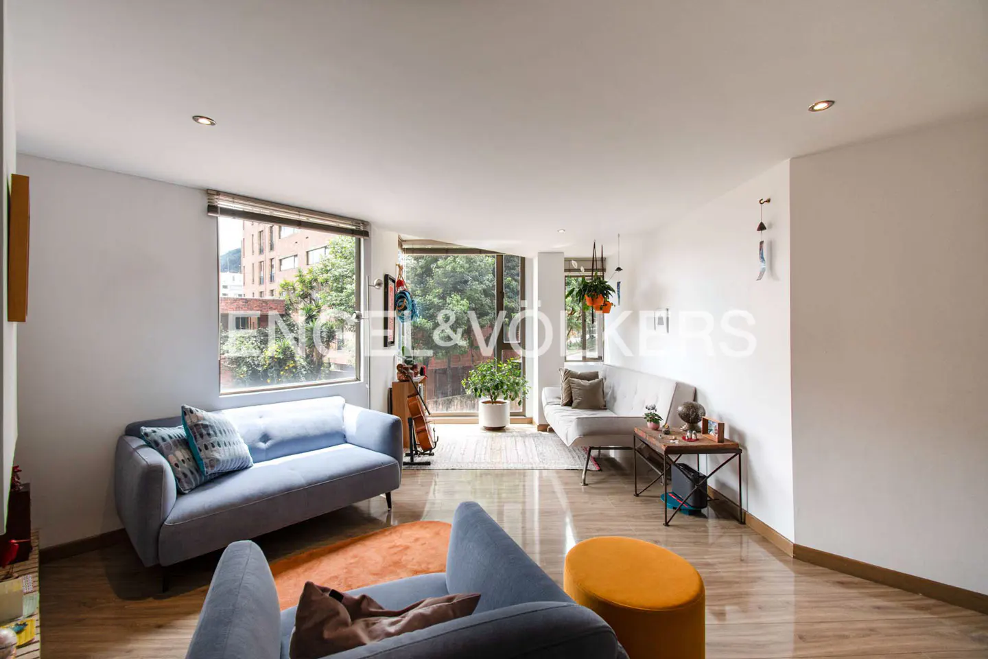 Bright living room with light blue sofas, wood floors, and large windows overlooking greenery. An orange rug and ottoman add warmth.