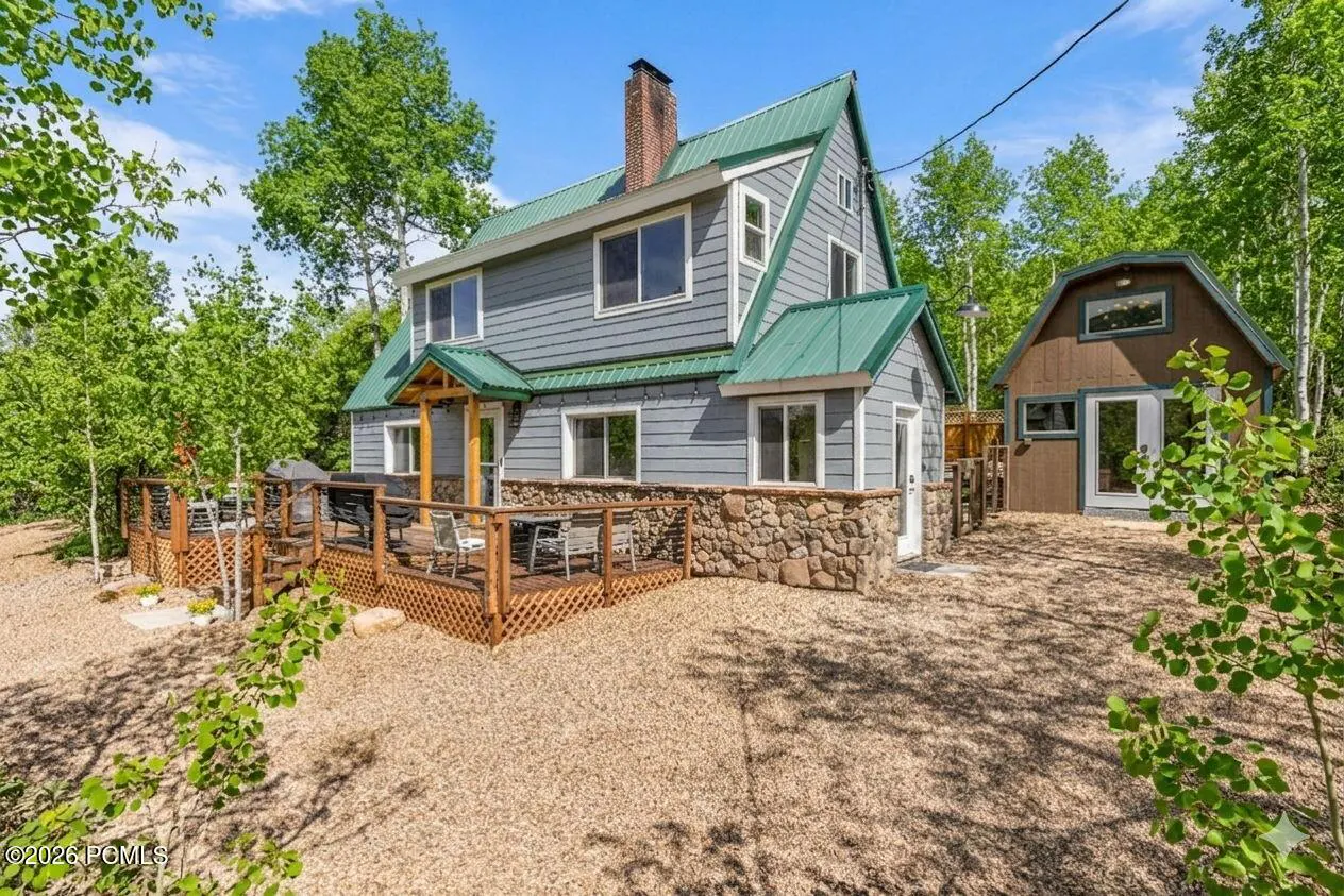 Two-story gray house with a green roof, stone foundation, and wooden deck, surrounded by trees and a gravel yard.