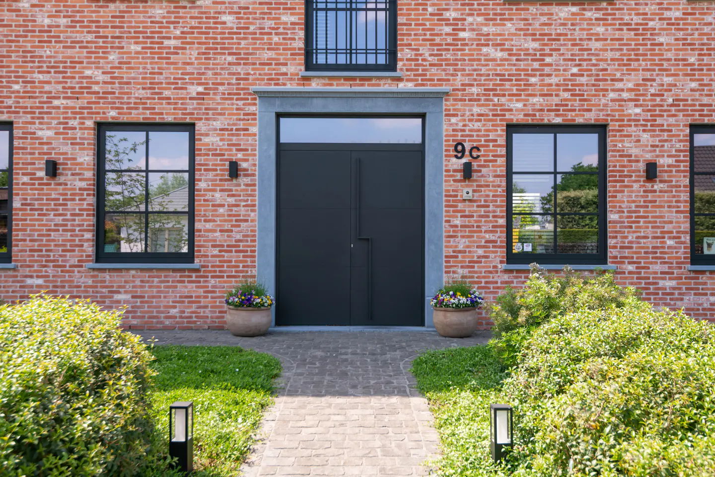 Brick house with a black double door, number 9c, flanked by windows and flower pots. A stone path leads to the entrance.