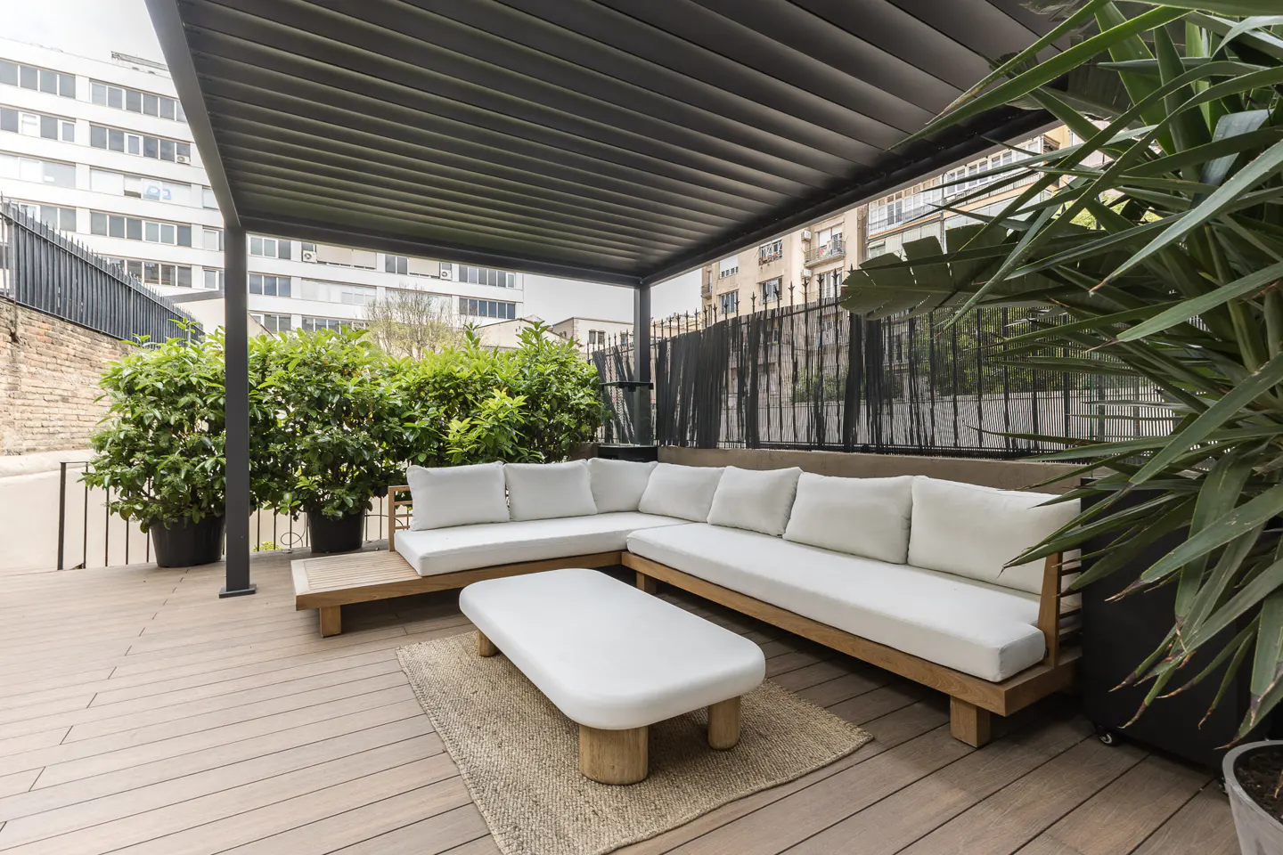 Outdoor patio with a wooden sectional sofa, white cushions, and a white coffee table on a jute rug. Green plants and a black pergola frame the space.