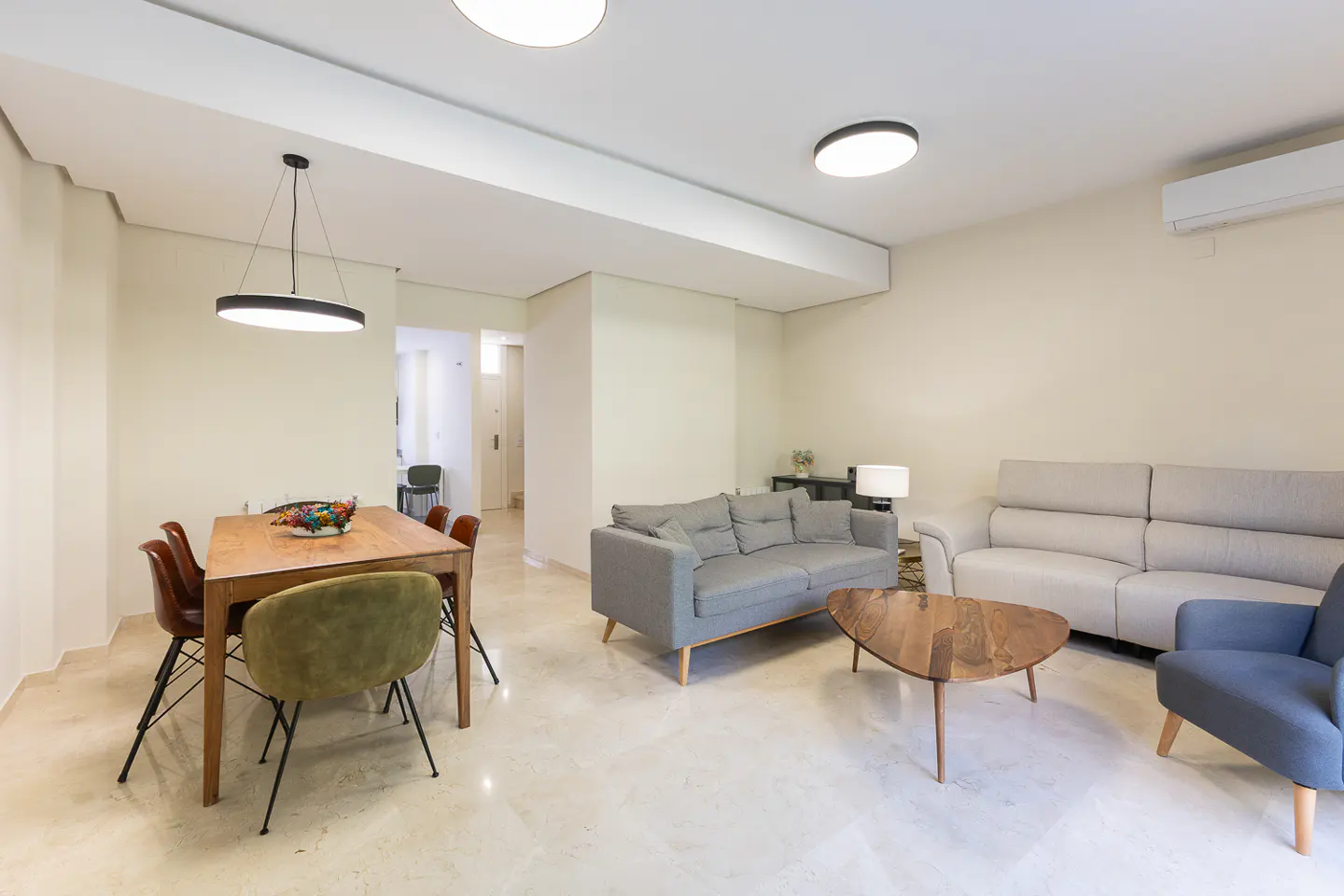 Modern open-plan living room with a wooden dining table, grey sofa, white couch, and blue armchair on marble floors.