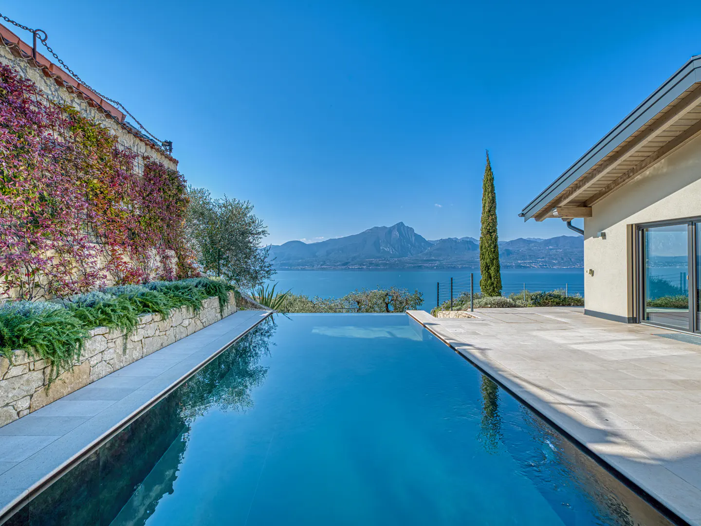 Infinity pool overlooking a blue lake and mountains under a clear sky. Stone wall with vines on the left, modern house on the right.