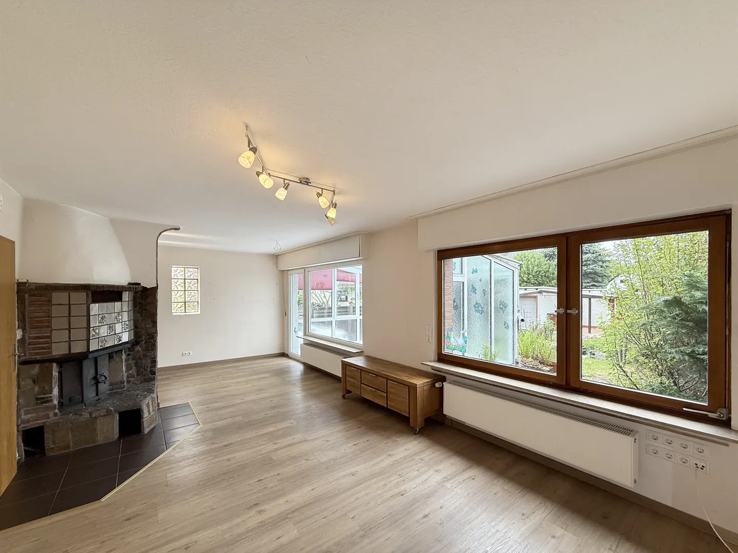 Bright living room with wood floors, a stone fireplace, and large windows overlooking a green yard. A wooden cabinet sits beneath the windows.