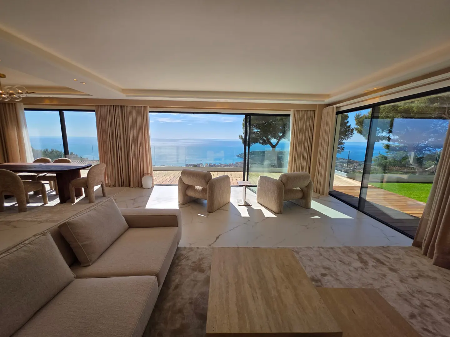 Bright living room with beige sofa, chairs, and dining table. Large windows offer ocean views. Marble floors reflect sunlight.