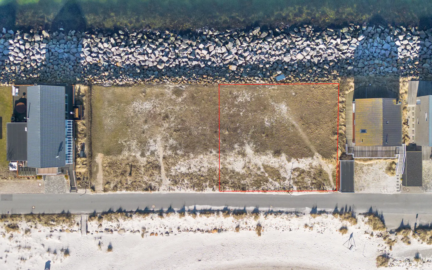 Aerial view of a vacant lot outlined in red, situated between houses, a road, and a sandy beach near a rocky shoreline.