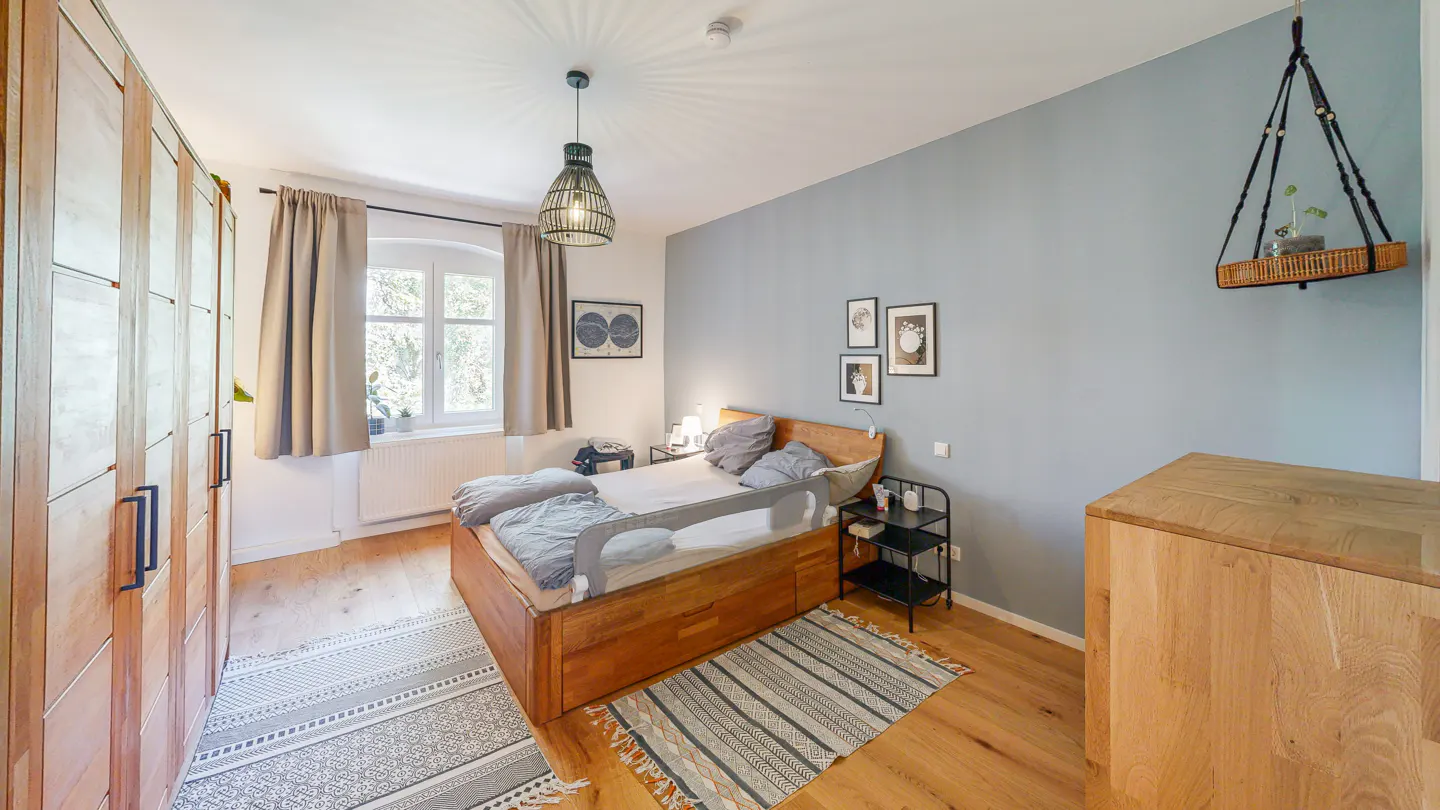 Bedroom with wooden bed, large wardrobe, blue accent wall, hanging plant shelf, and patterned rugs on hardwood floors.