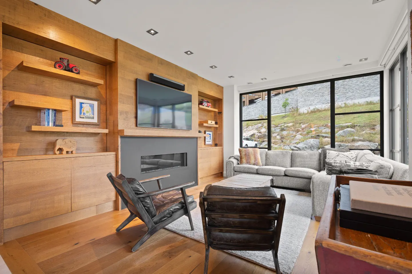Living room with wood paneling, a gray fireplace, and a large window with a view of a rocky hillside. Two leather chairs face a gray sofa.