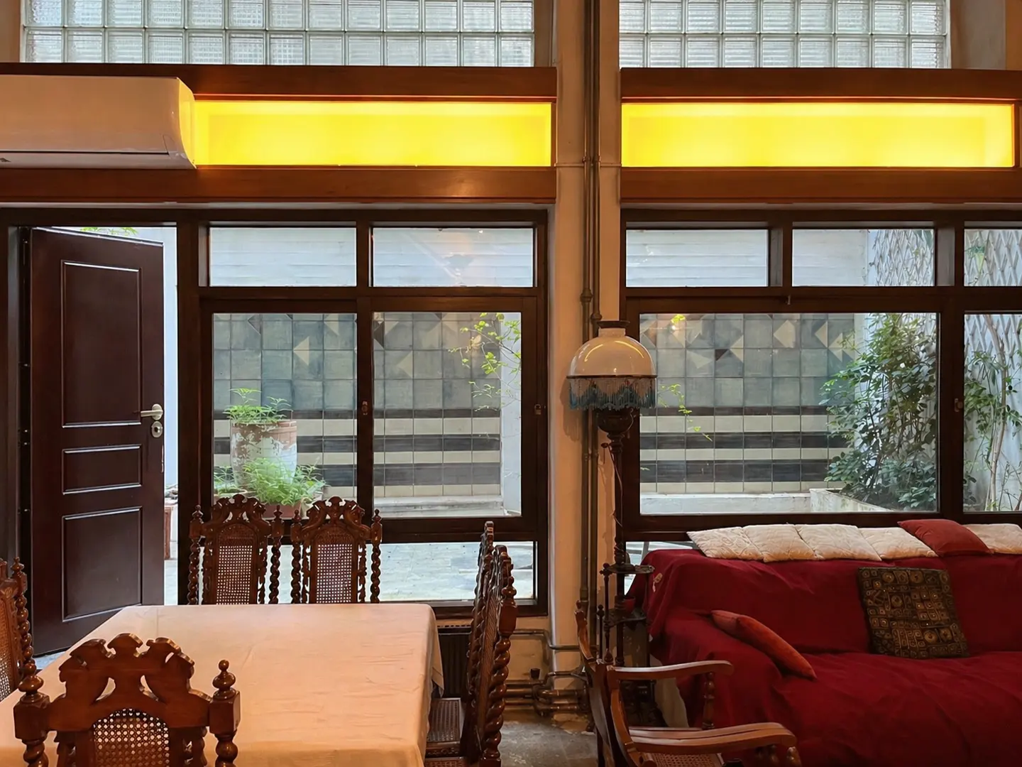 Interior view of a dining and living area with large windows. A table with chairs is on the left, and a red sofa is on the right.