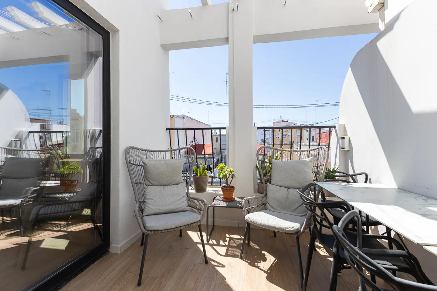 Balcony with two gray chairs, a small table with plants, and a white table with black chairs. City view in the background.
