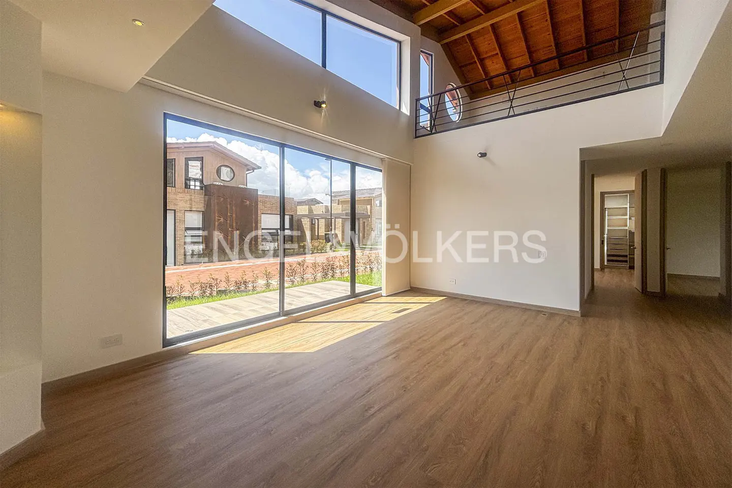 Bright, empty living room with wood floors, white walls, and large windows. A balcony with black railings overlooks the space. The Engel & Völkers logo is visible on the window.