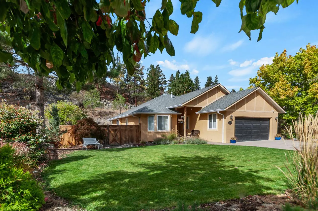 Tan single-story house with a gray roof, black garage door, and green lawn on a sunny day. A wooden fence and trees surround the property.