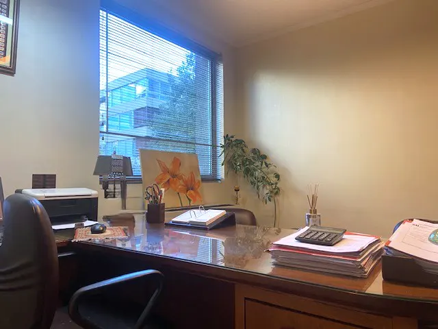 Office interior with a wooden desk, papers, calculator, and a window view of a building and trees.