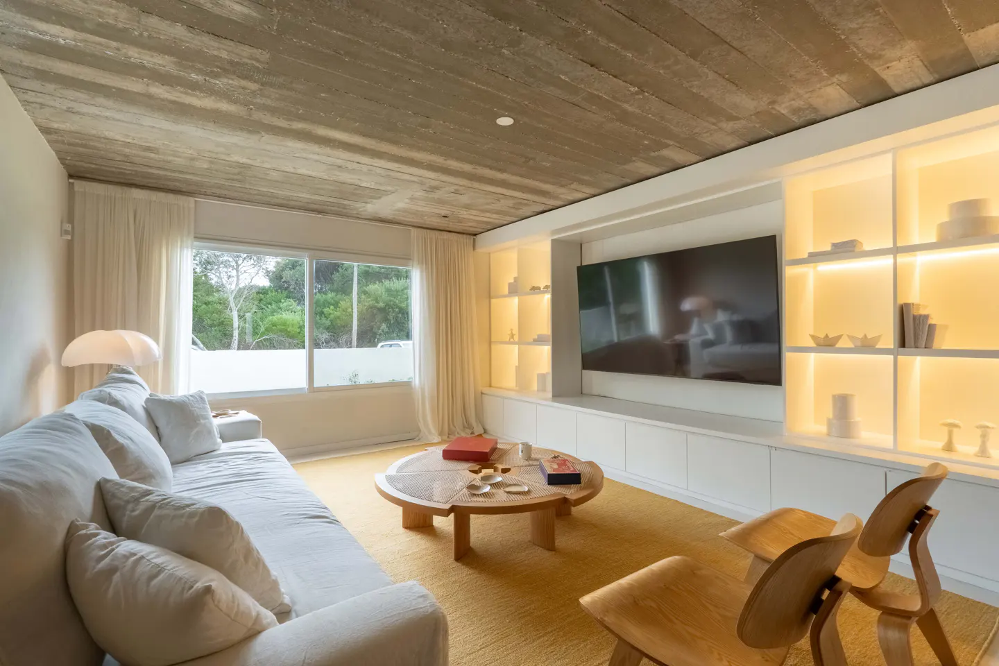 Bright living room with a white sofa, wooden chairs, and a round coffee table on a yellow rug. A large window and built-in shelves with a TV are visible.