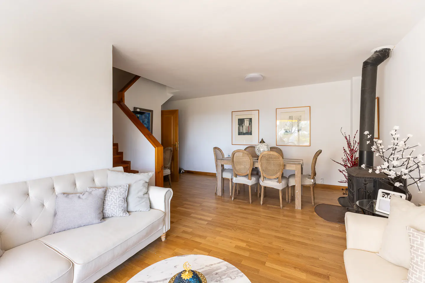 Bright living room with hardwood floors, white walls, and a staircase. A cream sofa sits near a marble table. A dining table and wood-burning stove are in the background.