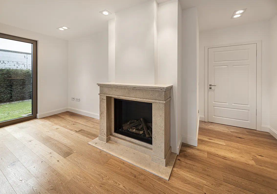 Bright, modern living room with hardwood floors, a stone fireplace, and a large window overlooking a green garden. A white door is visible in the background.