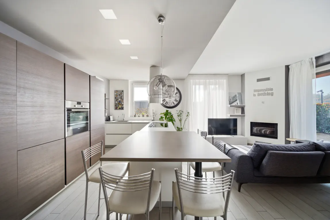 Modern open-plan kitchen and living area with wood cabinets, a central island, dining chairs, and a gray sofa.