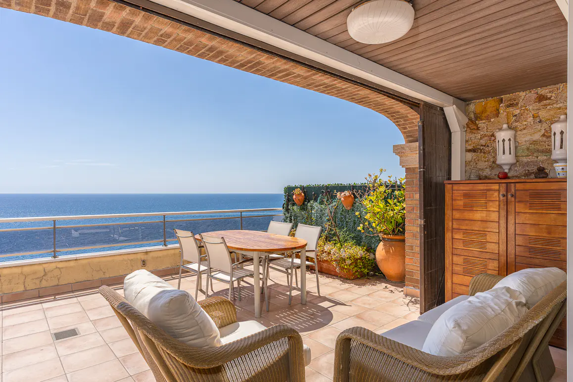 Outdoor patio with ocean view. Wicker chairs, table, and cabinet on tiled floor. Blue sky and ocean in background.
