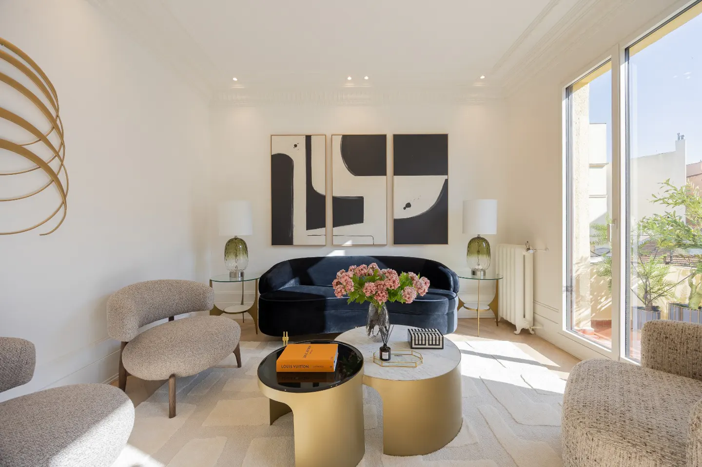Bright living room with a blue sofa, abstract art, and gold accents. Two textured chairs face a round coffee table with flowers. Large windows let in natural light.
