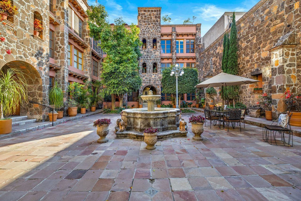 Courtyard view with stone fountain, potted plants, and outdoor seating. Buildings with stone walls and arched windows surround the space.