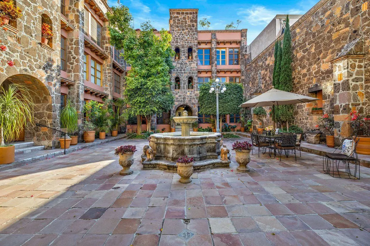 Courtyard view with stone fountain, potted plants, and outdoor seating. Buildings with stone walls and arched windows surround the space.