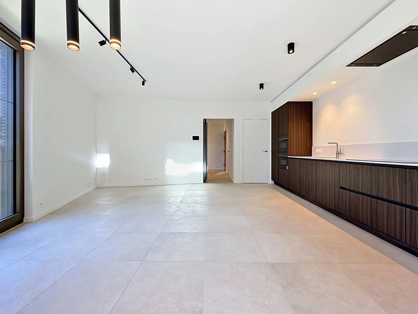 Bright, modern open-concept living space with white walls, tile floor, and dark wood kitchen cabinets. Black track lighting on the ceiling.