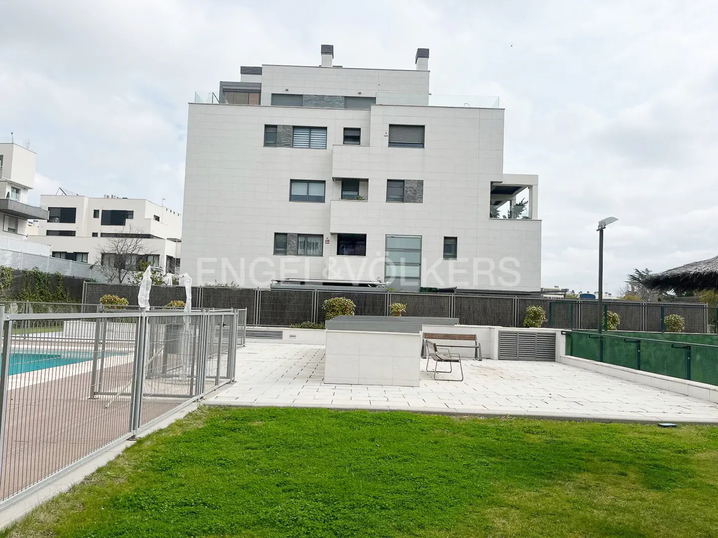 Modern white apartment building with a patio, swimming pool, and green lawn under a cloudy sky.