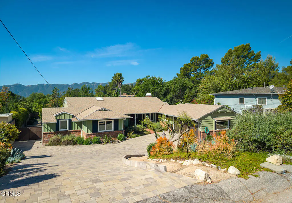 A green and brick house with a tan roof sits on a paved driveway under a clear blue sky. Trees and mountains are in the background.
