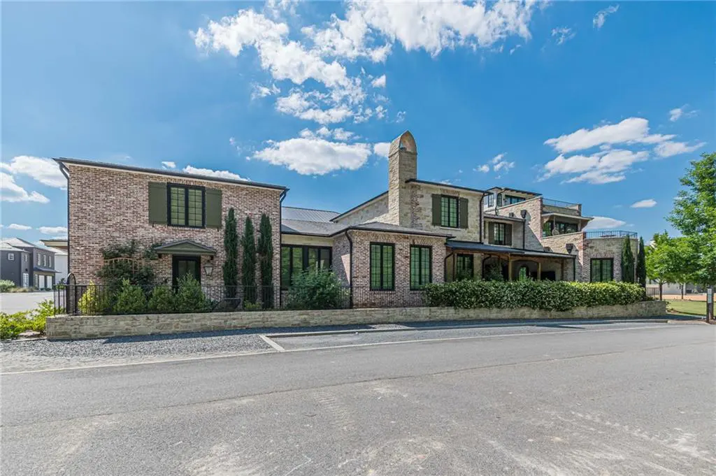 Large multi-story brick home with dark window frames, a stone chimney, and manicured hedges under a bright blue sky.
