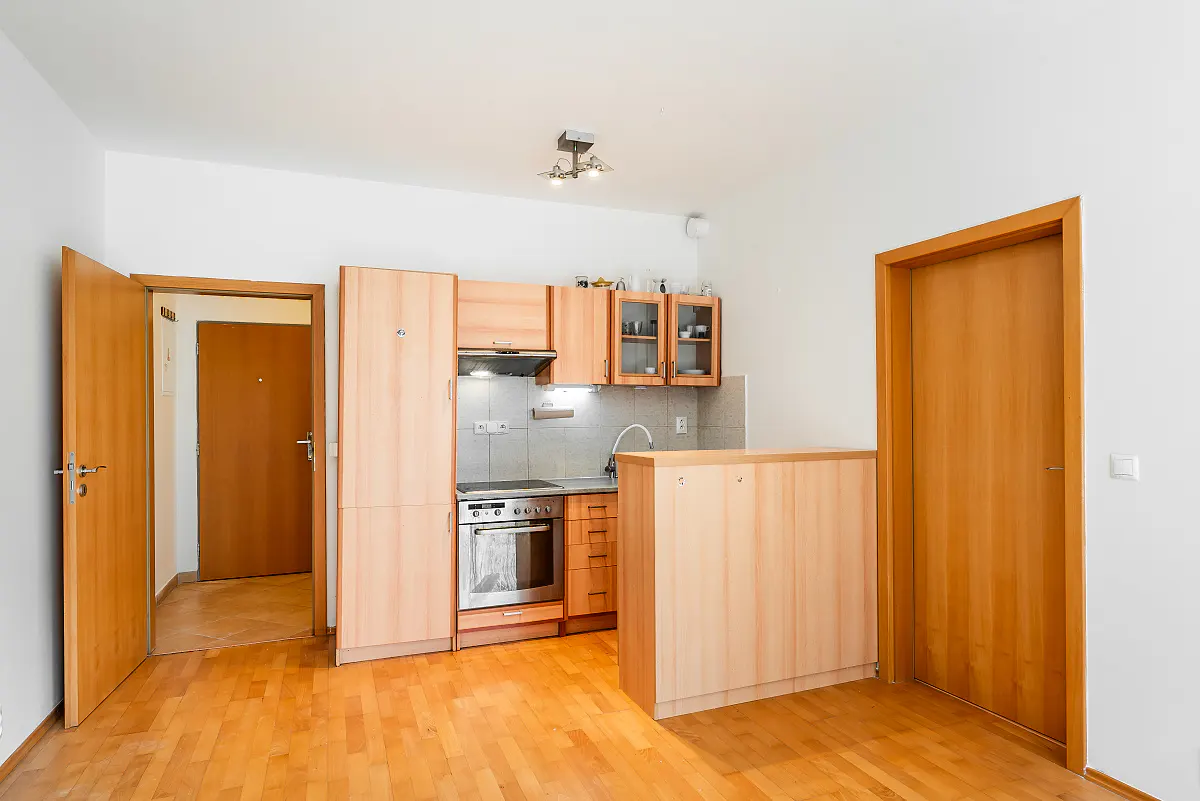 Bright apartment interior with light wood floors, white walls, and a compact kitchen area featuring wood cabinets and stainless steel appliances. Two wood doors are visible.